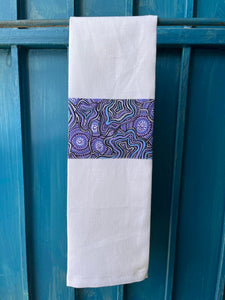 A white tea towel made from European linen and featuring the art work Meteors by Australian Indigenous artist Heather Kennedy. The tea towel is hanging on a deep blue coloured wall.