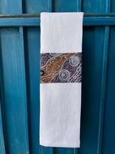 Load image into Gallery viewer, A white tea towel made from European linen and featuring the art work Bush Camp by Australian Indigenous artist Audrey Martin Napanangka. The tea towel is hanging on a deep blue coloured wall.
