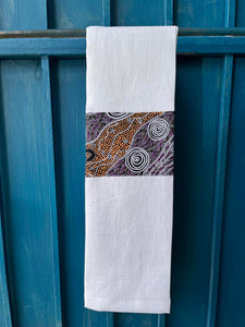 A white tea towel made from European linen and featuring the art work Bush Camp by Australian Indigenous artist Audrey Martin Napanangka. The tea towel is hanging on a deep blue coloured wall.