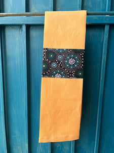 A yellow tea towel made from European linen and featuring the art work Gathering Bush Tucker by Australian Indigenous artist Gloria Doolan. The tea towel is hanging on a deep blue coloured wall.