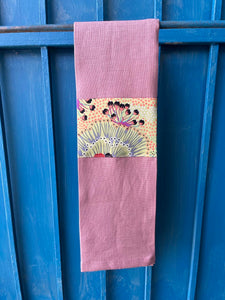 A rose pink tea towel made from European linen and featuring the art work Bush plum by Australian Indigenous artist Polly Wheeler. The tea towel is hanging on a deep blue coloured wall.