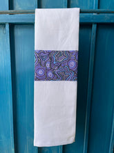 Load image into Gallery viewer, A white tea towel made from European linen and featuring the art work Meteors by Australian Indigenous artist Heather Kennedy. The tea towel is hanging on a deep blue coloured wall.