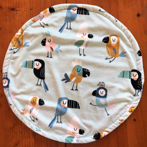 a round cat mat made with 3 layers of minky. Featuring toucan and parrot like birds in tan, black, blue and pink on a pale blue background